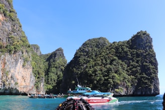 A picturesque seascape featuring towering limestone cliffs draped in lush greenery surrounded by clear blue water. Several boats, including a red speedboat and a blue ferry, are floating near the rocky shore.
