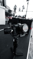 A digital camera mounted on a gimbal sits on a surface with a blurred outdoor background. The camera is equipped with a lens, and various controls and buttons are visible. The scene has a monochromatic tone with selective color in some details.