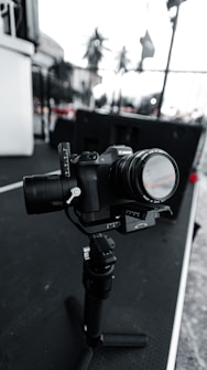 A digital camera mounted on a gimbal sits on a surface with a blurred outdoor background. The camera is equipped with a lens, and various controls and buttons are visible. The scene has a monochromatic tone with selective color in some details.