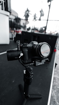 A digital camera mounted on a gimbal sits on a surface with a blurred outdoor background. The camera is equipped with a lens, and various controls and buttons are visible. The scene has a monochromatic tone with selective color in some details.