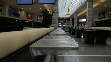 A spacious shopping mall food court with empty tables covered in protective plastic. The setting features modern architecture with a high ceiling and large windows that allow natural light to enter. Signs for various eateries are visible, along with decorative plants and a contemporary art installation hanging from the ceiling.
