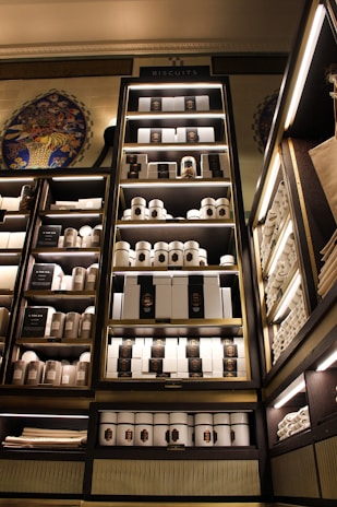 A well-organized display shelf filled with neatly arranged biscuit tins and boxes. The shelves are illuminated, emphasizing the elegant packaging of the products. The setting appears to be a high-end store with decorative elements like intricate wall designs. The colors and lighting create a luxurious atmosphere.
