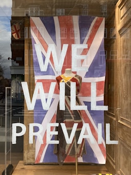 A storefront window display features the large text 'WE WILL PREVAIL' painted in white over a Union Jack flag backdrop. In front of the flag, a dress on a mannequin incorporates similar colors and patterns, aligning with the British theme. Reflections of nearby buildings are visible on the glass.