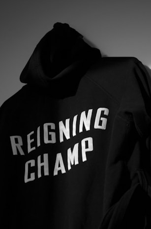 A black hoodie with the words 'REIGNING CHAMP' printed in bold white letters on the back. The fabric appears soft and heavy, casting a shadow in a dimly lit environment.