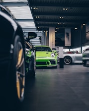A vibrant green sports car is prominently displayed in the center of an upscale car showroom. The showroom features a modern design with high ceilings and extensive use of glass, allowing natural light to illuminate the vehicles. Additional sports cars in various colors are visible, suggesting a high-end collection.