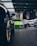 A vibrant green sports car is prominently displayed in the center of an upscale car showroom. The showroom features a modern design with high ceilings and extensive use of glass, allowing natural light to illuminate the vehicles. Additional sports cars in various colors are visible, suggesting a high-end collection.