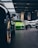 A vibrant green sports car is prominently displayed in the center of an upscale car showroom. The showroom features a modern design with high ceilings and extensive use of glass, allowing natural light to illuminate the vehicles. Additional sports cars in various colors are visible, suggesting a high-end collection.