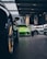 A vibrant green sports car is prominently displayed in the center of an upscale car showroom. The showroom features a modern design with high ceilings and extensive use of glass, allowing natural light to illuminate the vehicles. Additional sports cars in various colors are visible, suggesting a high-end collection.