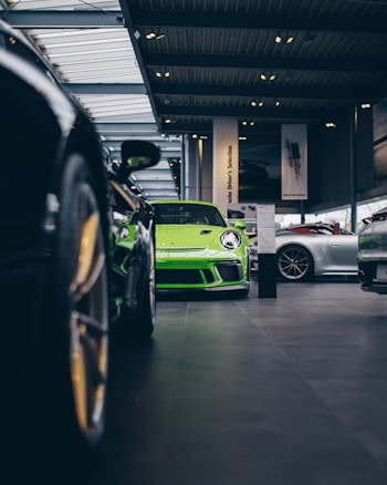 A vibrant green sports car is prominently displayed in the center of an upscale car showroom. The showroom features a modern design with high ceilings and extensive use of glass, allowing natural light to illuminate the vehicles. Additional sports cars in various colors are visible, suggesting a high-end collection.