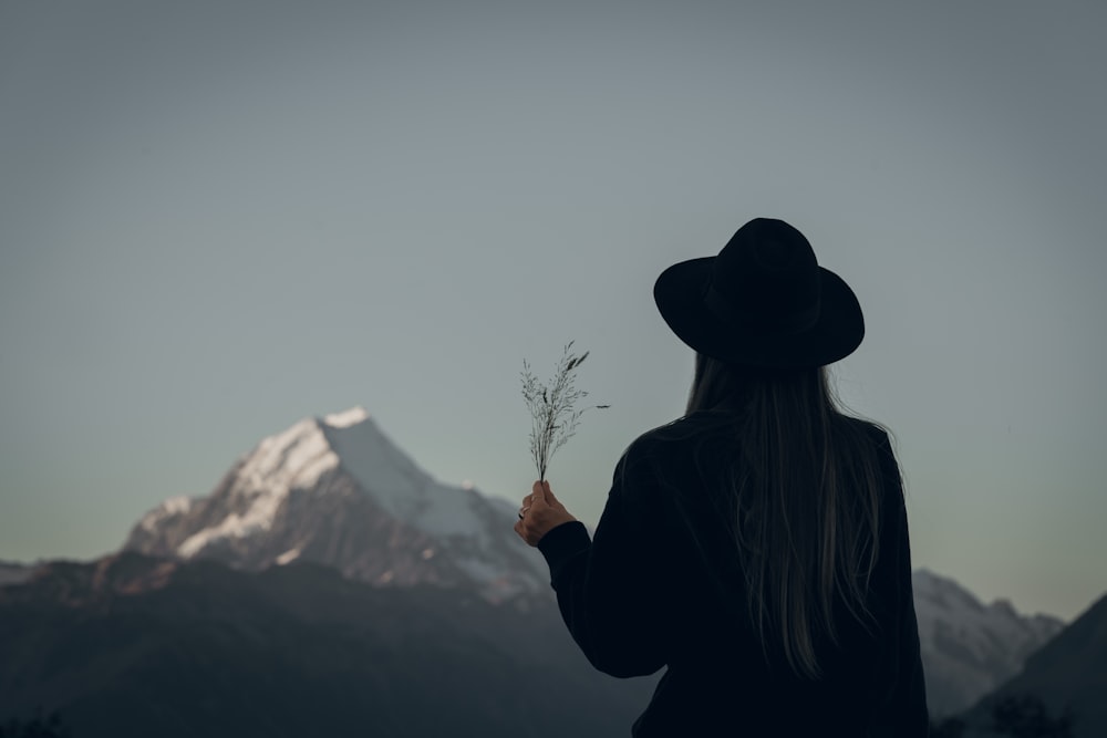 검은 재킷과 갈색 모자를 쓴 여자가 낮에 눈 덮인 산 근처에 서 있다