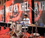 A man is playing a drum set on a stage. The backdrop displays the text 'WAQT KA KHEL' in bold white letters over a colorful design. Multiple microphones are positioned around the drum set, indicating a live performance or recording setup. The stage is covered with a red carpet, and there are speakers and other equipment visible in the background.