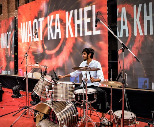A man is playing a drum set on a stage. The backdrop displays the text 'WAQT KA KHEL' in bold white letters over a colorful design. Multiple microphones are positioned around the drum set, indicating a live performance or recording setup. The stage is covered with a red carpet, and there are speakers and other equipment visible in the background.