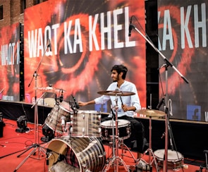 A man is playing a drum set on a stage. The backdrop displays the text 'WAQT KA KHEL' in bold white letters over a colorful design. Multiple microphones are positioned around the drum set, indicating a live performance or recording setup. The stage is covered with a red carpet, and there are speakers and other equipment visible in the background.