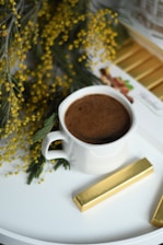 A white cup filled with a frothy brown beverage is placed on a white surface. Surrounding the cup are sprigs of green foliage adorned with small yellow flowers. Beside the cup, there is a gold-wrapped chocolate bar, complementing the color scheme.