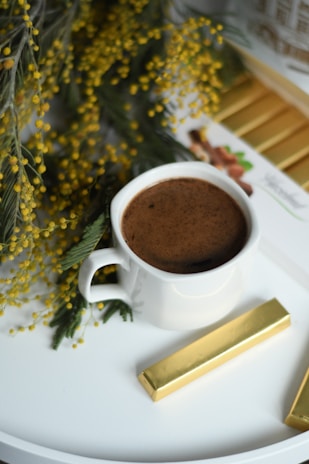 A white cup filled with a frothy brown beverage is placed on a white surface. Surrounding the cup are sprigs of green foliage adorned with small yellow flowers. Beside the cup, there is a gold-wrapped chocolate bar, complementing the color scheme.