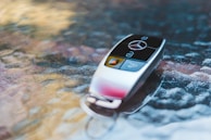 A sleek car key with a metallic finish rests on a textured glass surface. The key features a logo and prominent buttons.