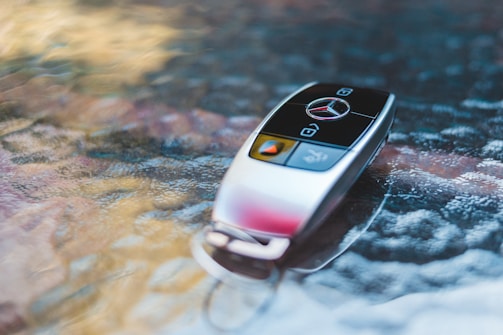 A sleek car key with a metallic finish rests on a textured glass surface. The key features a logo and prominent buttons.