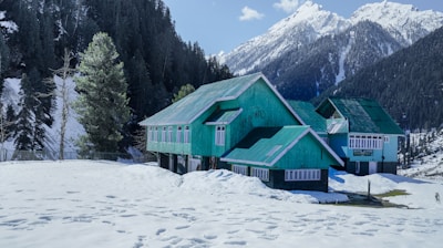 green wooden house on snow covered ground near green trees and mountain during daytime