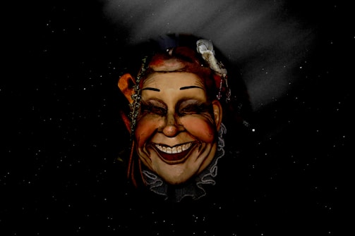 A realistic and expressive theatrical mask of a smiling face with exaggerated features, including high-arched eyebrows, red cheeks, and a wide grin, is depicted against a dark, starry background. The mask has accessories, such as decorative headpieces and what appears to be ribbons or strings.