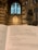 An open book displaying a religious text inside a cathedral, with a focus on the Lord’s Prayer. The background features ornate architecture and towering stained glass windows in a gothic style.
