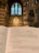 An open book displaying a religious text inside a cathedral, with a focus on the Lord’s Prayer. The background features ornate architecture and towering stained glass windows in a gothic style.