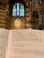 An open book displaying a religious text inside a cathedral, with a focus on the Lord’s Prayer. The background features ornate architecture and towering stained glass windows in a gothic style.