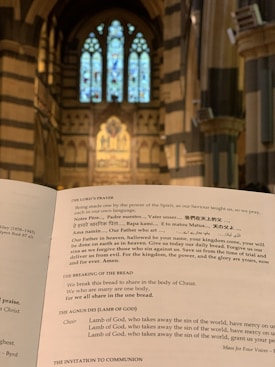An open book displaying a religious text inside a cathedral, with a focus on the Lord’s Prayer. The background features ornate architecture and towering stained glass windows in a gothic style.