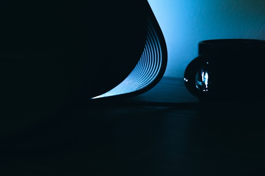 A close-up photograph of a book with light illuminating its curved pages, creating a dramatic contrast against the dark surroundings. The focus is on the page ridges, with a soft blue light casting subtle shadows. In the background, a blurred dark object adds depth to the composition.