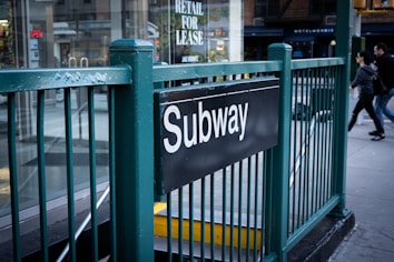 A subway entrance marked with a black sign featuring the word 'Subway' in white letters. The entrance is surrounded by green metal railings, and people are walking on the adjacent sidewalk. A glass storefront with various signage, including one for lease, is visible in the background.