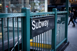 A subway entrance marked with a black sign featuring the word 'Subway' in white letters. The entrance is surrounded by green metal railings, and people are walking on the adjacent sidewalk. A glass storefront with various signage, including one for lease, is visible in the background.