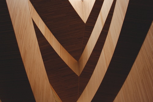 A geometric design featuring large triangular wooden panels creating a symmetrical pattern. The panels are arranged in multiple layers, casting shadows and creating a visual illusion of depth and movement.