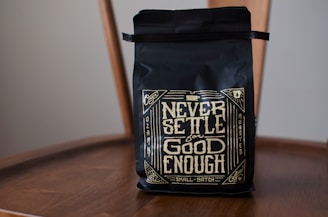 A black coffee bag with decorative golden lettering displaying the phrase 'Never Settle for Good Enough'. The bag is placed on a wooden chair, and the design includes intricate patterns and vintage motifs.