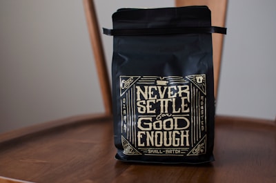A black coffee bag with decorative golden lettering displaying the phrase 'Never Settle for Good Enough'. The bag is placed on a wooden chair, and the design includes intricate patterns and vintage motifs.
