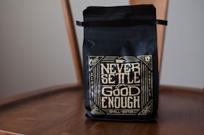 A black coffee bag with decorative golden lettering displaying the phrase 'Never Settle for Good Enough'. The bag is placed on a wooden chair, and the design includes intricate patterns and vintage motifs.