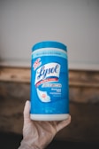A container of Lysol Advanced disinfecting wipes is held by a hand wearing a glove. The container is predominantly blue and white, featuring labels that emphasize its disinfecting power and ability to kill viruses and bacteria.