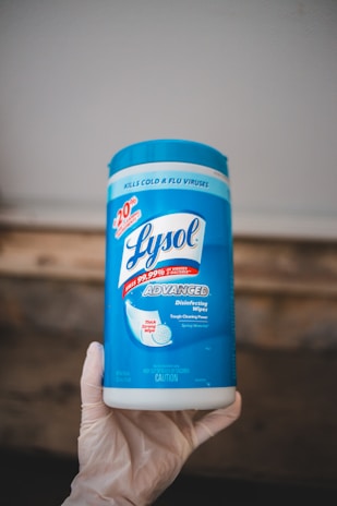 A container of Lysol Advanced disinfecting wipes is held by a hand wearing a glove. The container is predominantly blue and white, featuring labels that emphasize its disinfecting power and ability to kill viruses and bacteria.
