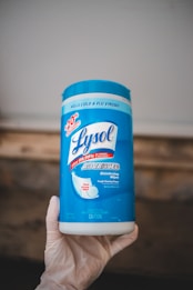 A container of Lysol Advanced disinfecting wipes is held by a hand wearing a glove. The container is predominantly blue and white, featuring labels that emphasize its disinfecting power and ability to kill viruses and bacteria.