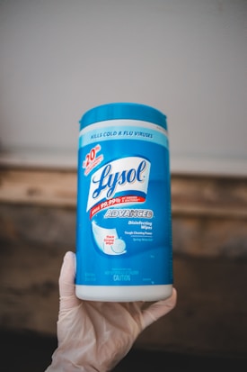 A container of Lysol Advanced disinfecting wipes is held by a hand wearing a glove. The container is predominantly blue and white, featuring labels that emphasize its disinfecting power and ability to kill viruses and bacteria.