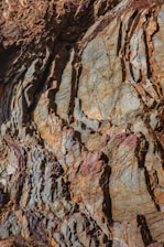 A close-up view of a textured rock surface with a mix of colors and intricate patterns. The rock face displays layers of different hues, including browns, grays, and hints of reddish tones. The surface is rugged, with visible striations and cracks, providing a detailed look at geological formations.