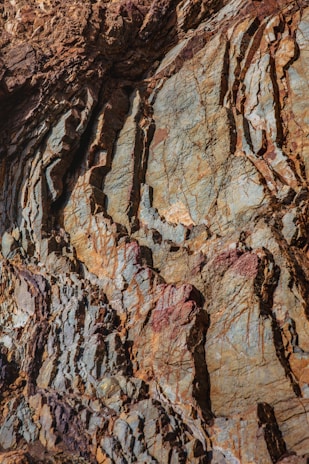 A close-up view of a textured rock surface with a mix of colors and intricate patterns. The rock face displays layers of different hues, including browns, grays, and hints of reddish tones. The surface is rugged, with visible striations and cracks, providing a detailed look at geological formations.