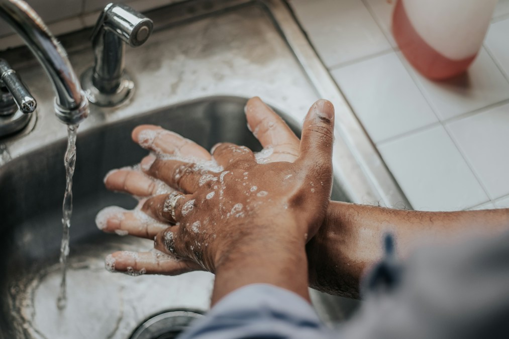 Des mains couvertes de mousse de savon se lavent sous le robinet d’un évier en inox