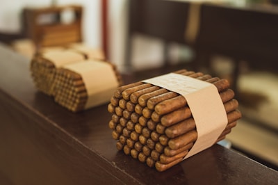 Bundles of cigars tied with beige bands rest on a wooden surface, with a blurred background. The cigars are neatly stacked and wrapped, giving a sense of careful arrangement.