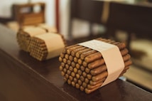 Bundles of cigars tied with beige bands rest on a wooden surface, with a blurred background. The cigars are neatly stacked and wrapped, giving a sense of careful arrangement.