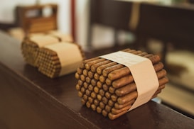 Bundles of cigars tied with beige bands rest on a wooden surface, with a blurred background. The cigars are neatly stacked and wrapped, giving a sense of careful arrangement.