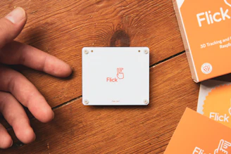 A white Flick HAT device is placed on a wooden table. To the left is a hand, slightly out of focus, reaching towards the device. On the right, there are orange boxes with 'Flick' branding and text indicating 3D tracking and gestures for Raspberry Pi.