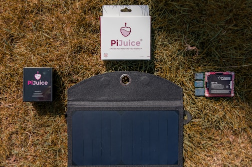 Various components of a PiJuice kit, including a solar panel, battery pack, and electronic boards, are laid out on dry grass.