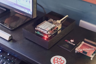 A small computing setup featuring a Raspberry Pi with a PiJuice HAT sits atop a black rectangular case on a desk. A monitor is positioned nearby, partially visible. There are various accessories, including PiJuice components and a Raspberry Pi-themed sticker.