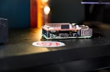 A small single-board computer is placed on a dark surface, accompanied by a sticker with a red and white logo. The computer features a series of ports and connectors, along with a heat sink on top, indicating its compact and functional design. The background is blurred with a warm glow.