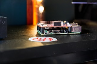 A small single-board computer is placed on a dark surface, accompanied by a sticker with a red and white logo. The computer features a series of ports and connectors, along with a heat sink on top, indicating its compact and functional design. The background is blurred with a warm glow.
