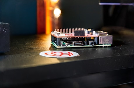 A small single-board computer is placed on a dark surface, accompanied by a sticker with a red and white logo. The computer features a series of ports and connectors, along with a heat sink on top, indicating its compact and functional design. The background is blurred with a warm glow.